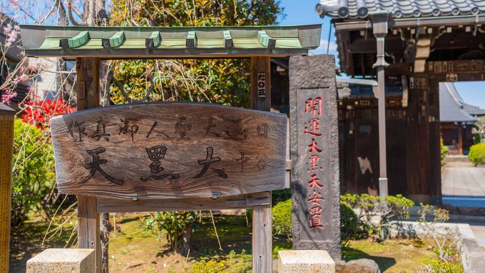 Ein Holzschild mit japanischen Schriftzeichen, eine steinerne Säule mit weiteren Schriftzeichen und ein traditionelles Gebäude im Hintergrund.