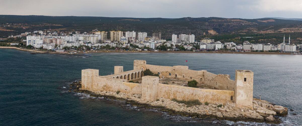 A coastal city with a historic fort on a rocky outcrop, modern buildings, and a hilly backdrop.
