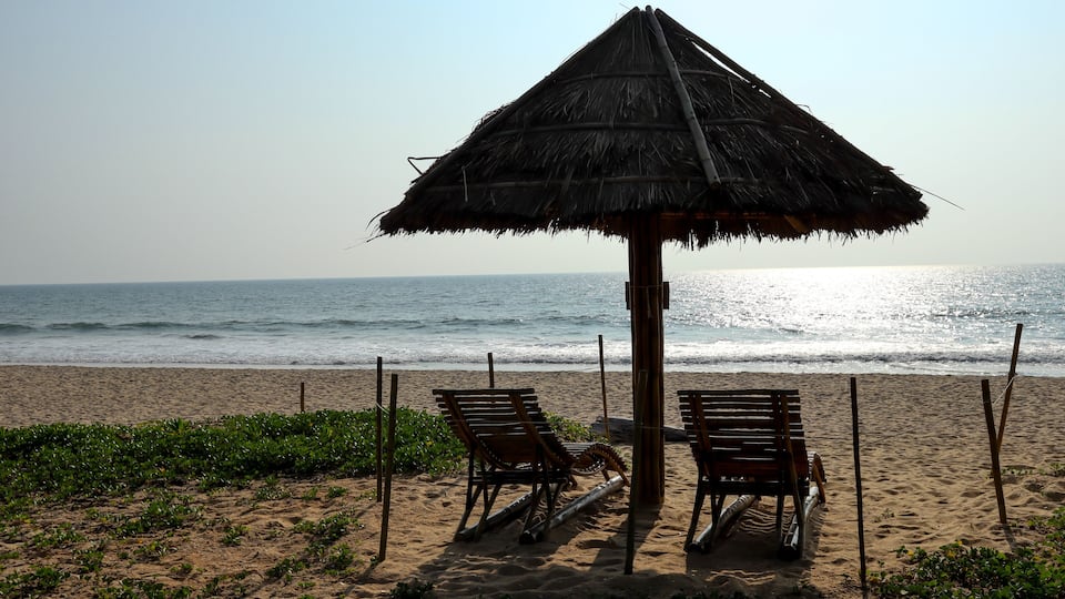 A beach with two wooden lounge chairs under a thatched umbrella, a sandy shore, and the ocean in the background.
