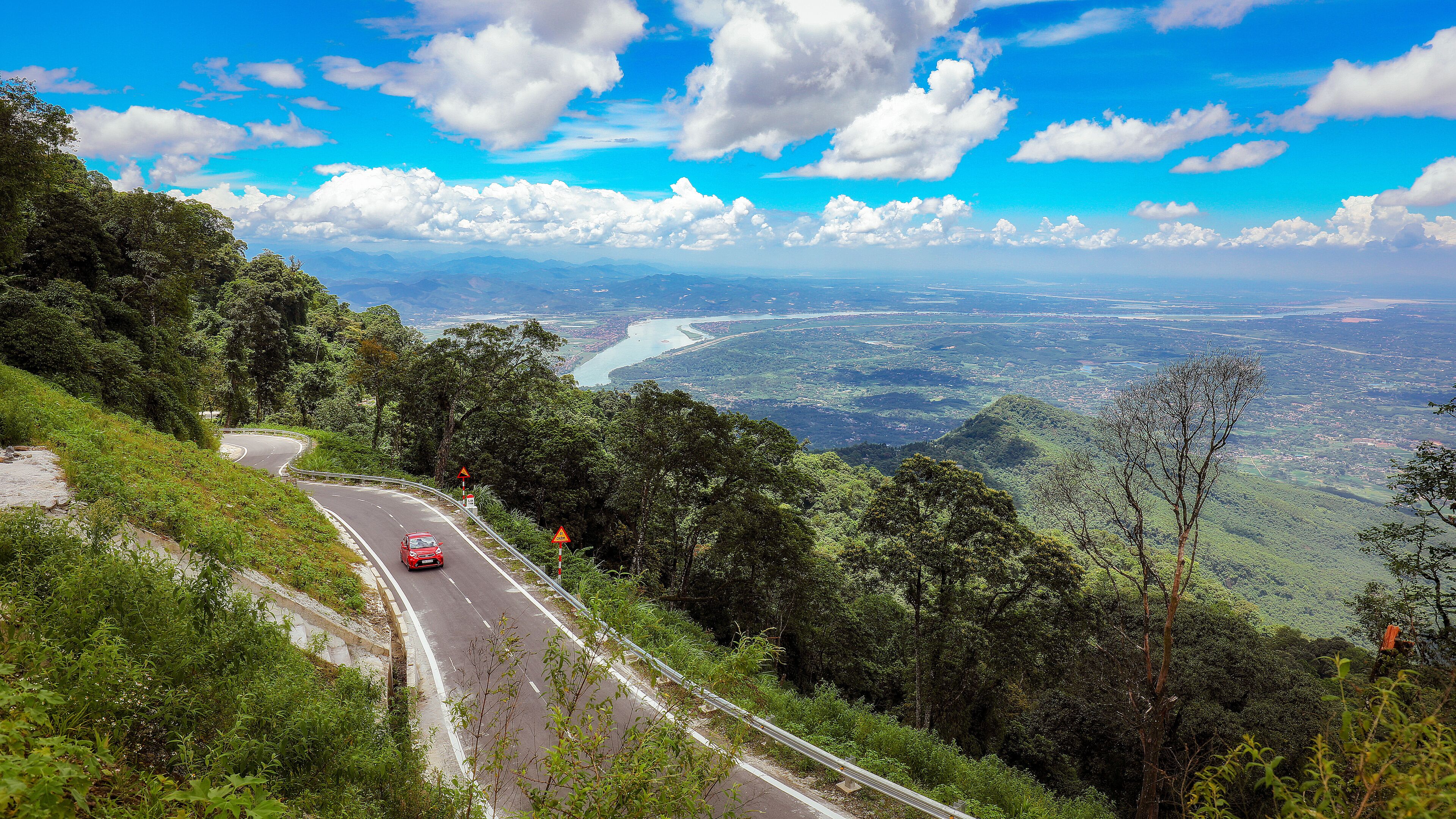 A winding road through a mountainous landscape with a river and city in the distance.