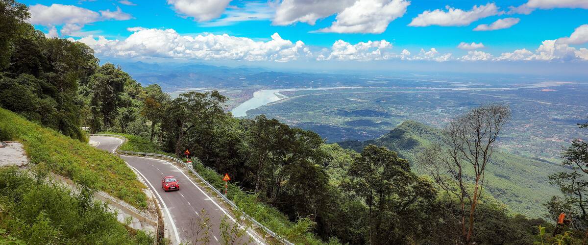 A winding road through a mountainous landscape with a river and city in the distance.