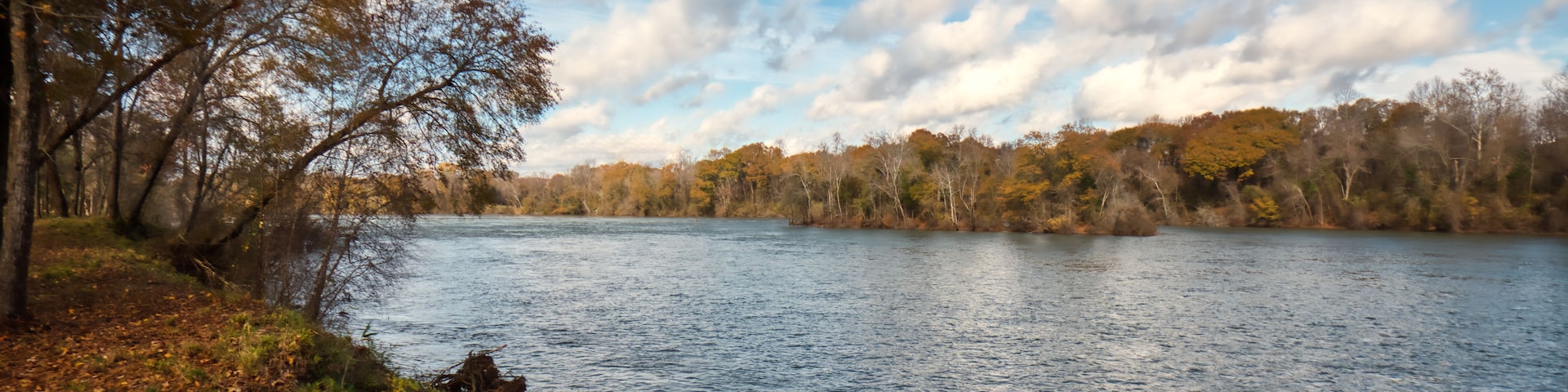 A calm lake surrounded by trees with autumn foliage, clear skies, and a few clouds.