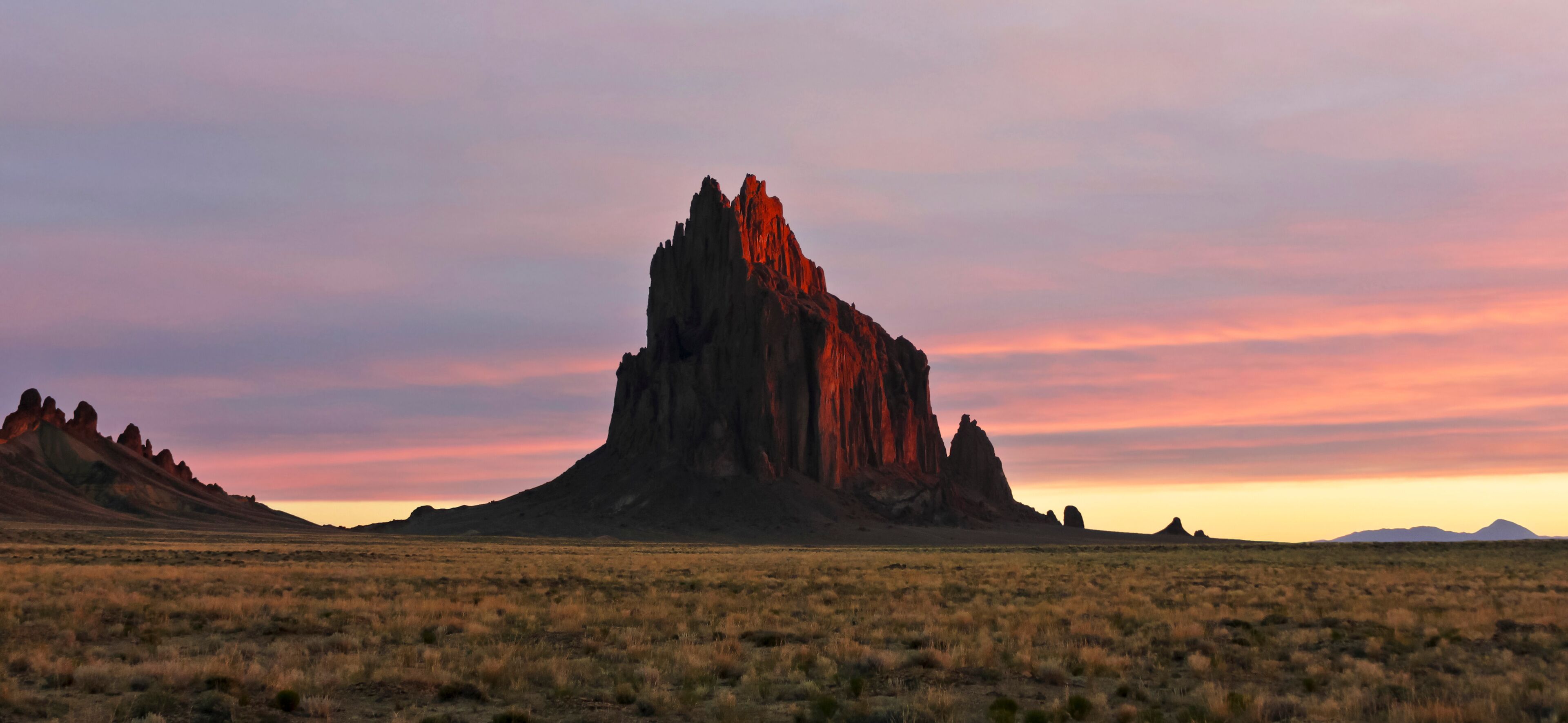 A desert landscape with a prominent rock formation in the center and distant mountains.