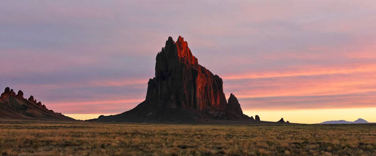 A desert landscape with a prominent rock formation in the center and distant mountains.
