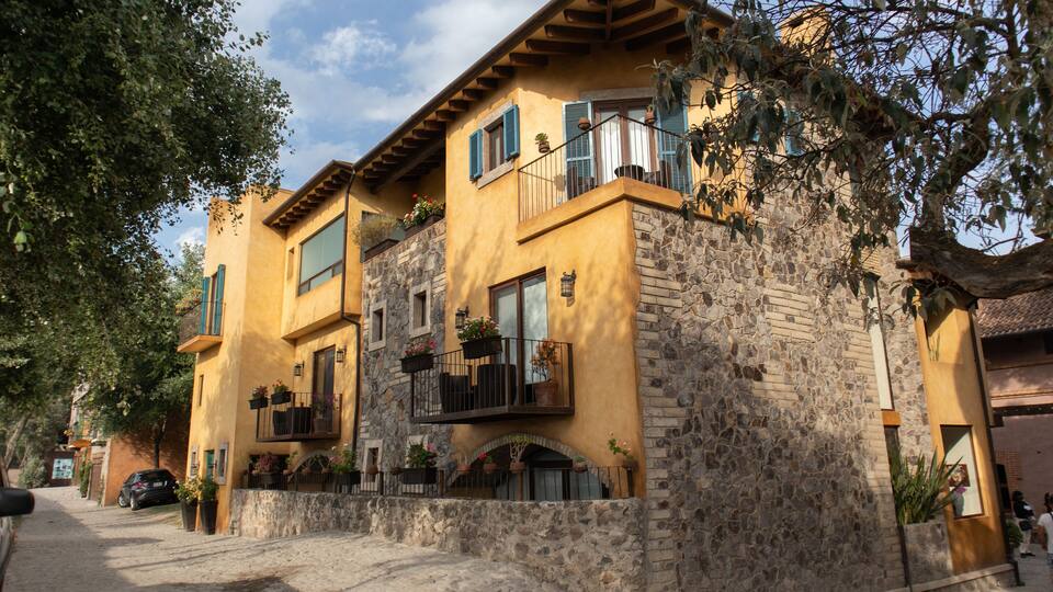 A yellow building with stone walls, balconies with potted plants, and a car parked in front.