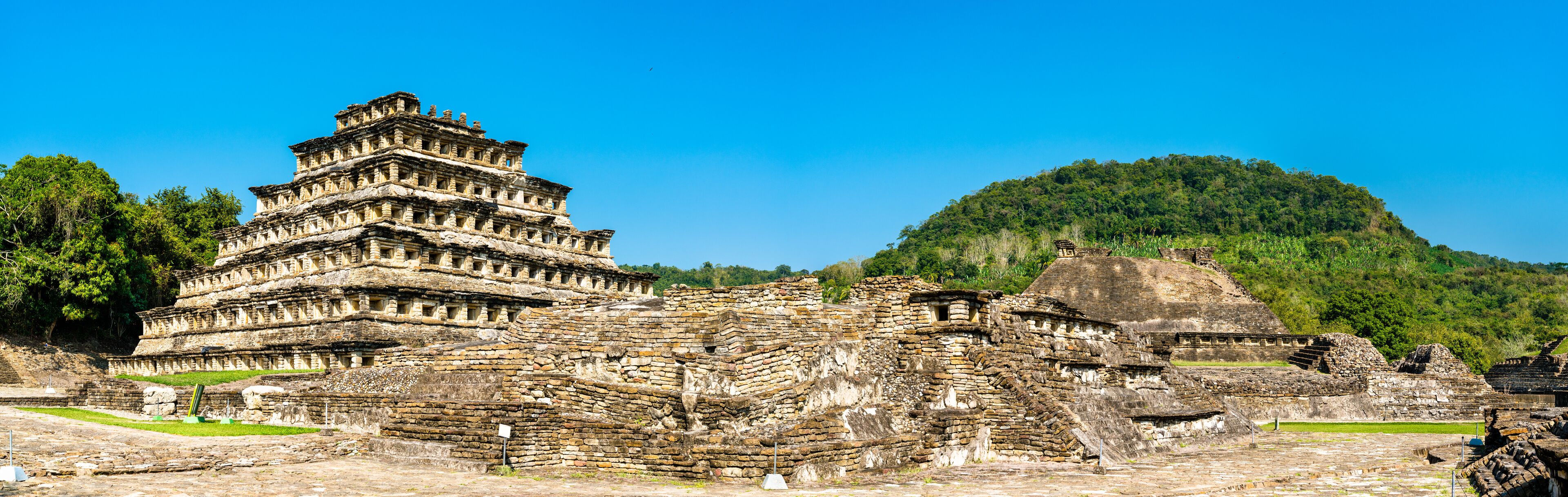An ancient pyramid-like structure with multiple tiers, surrounded by ruins and a lush green hill in the background.