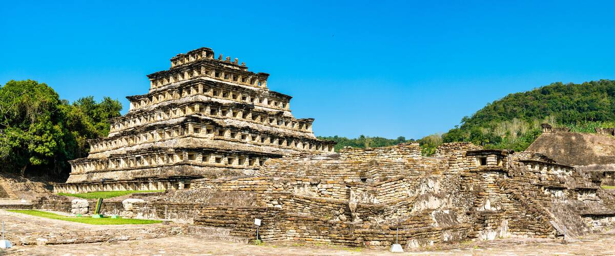 An ancient pyramid-like structure with multiple tiers, surrounded by ruins and a lush green hill in the background.