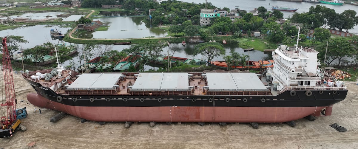 A large cargo ship docked at a port with a river and industrial buildings in the background.