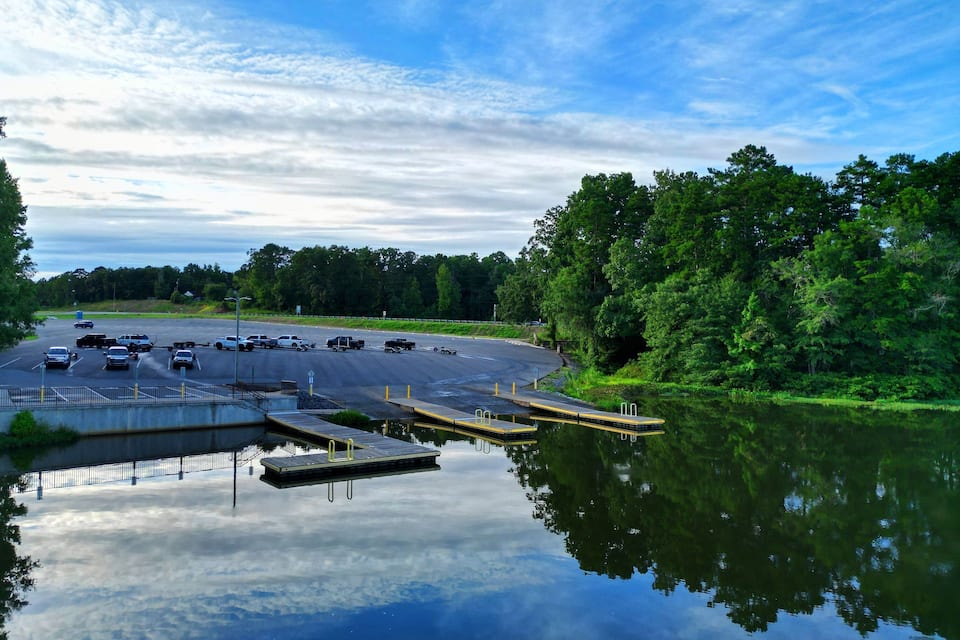 A parking lot with vehicles, a dock extending into a calm lake, and dense trees in the background.
