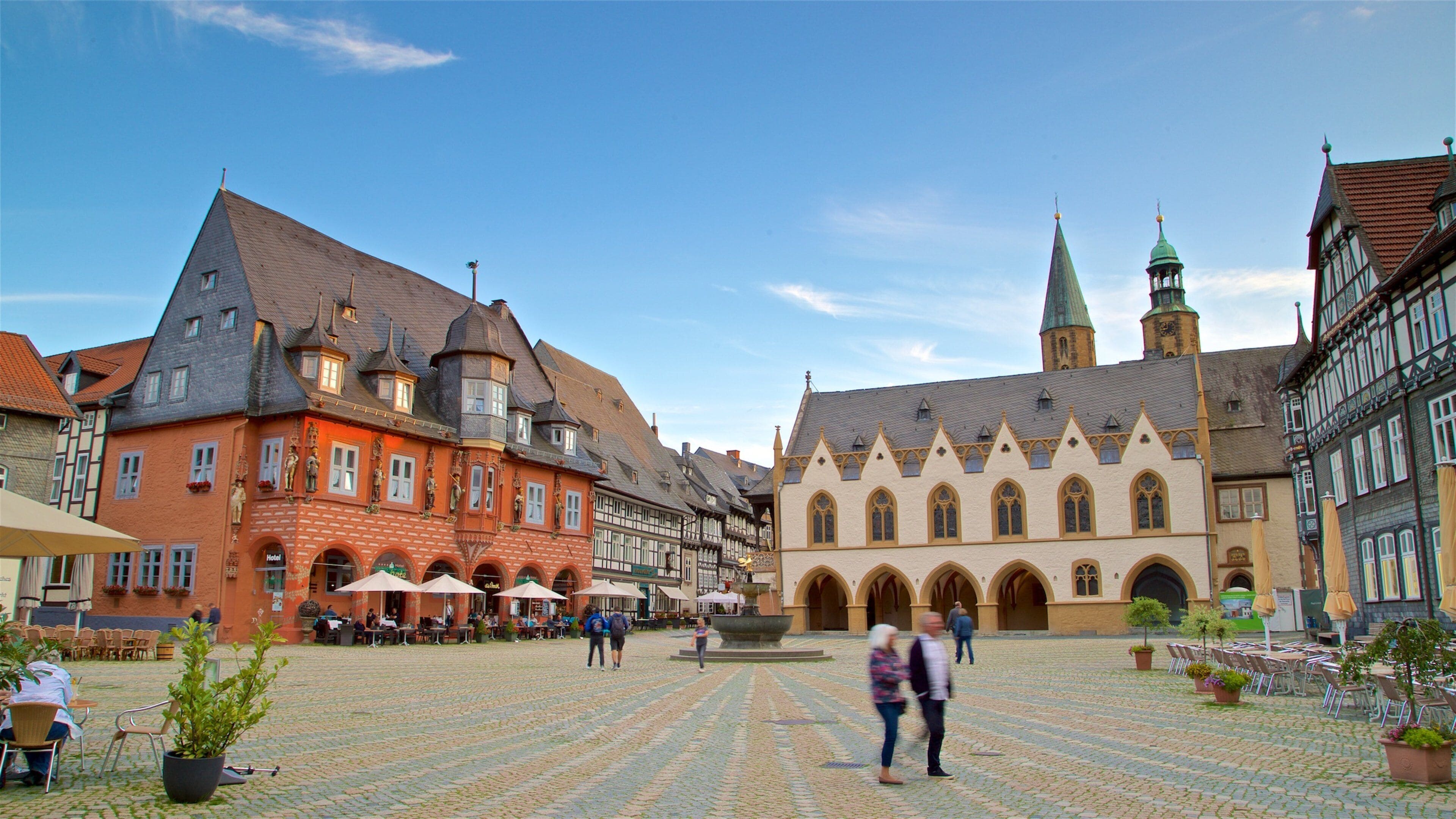 A cobblestone square with historic buildings, a church tower, and outdoor seating areas.