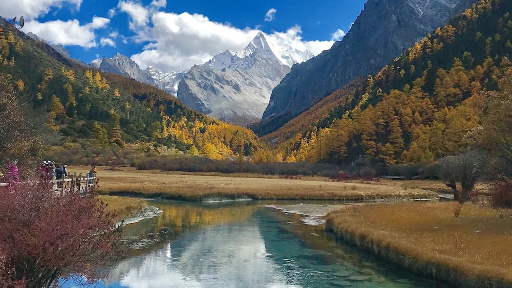 A clear mountain lake surrounded by autumn-colored trees and snow-capped peaks.