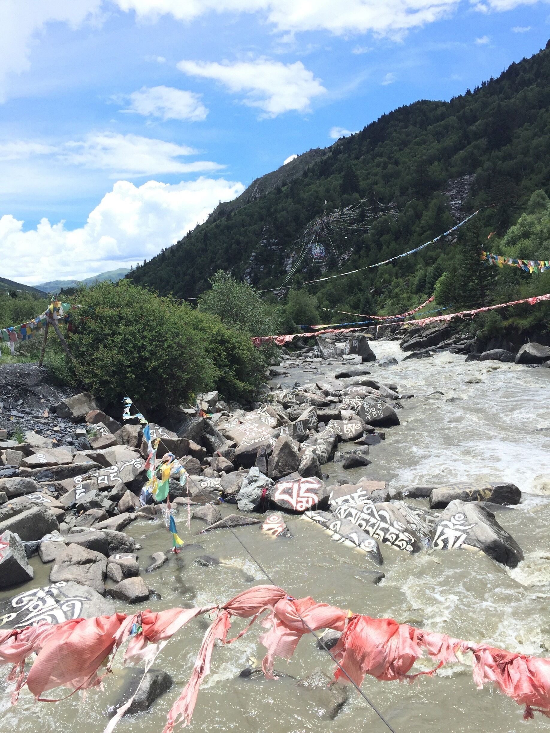 A river with rocks and a person crossing. There are colorful flags and a mountainous landscape in the background.