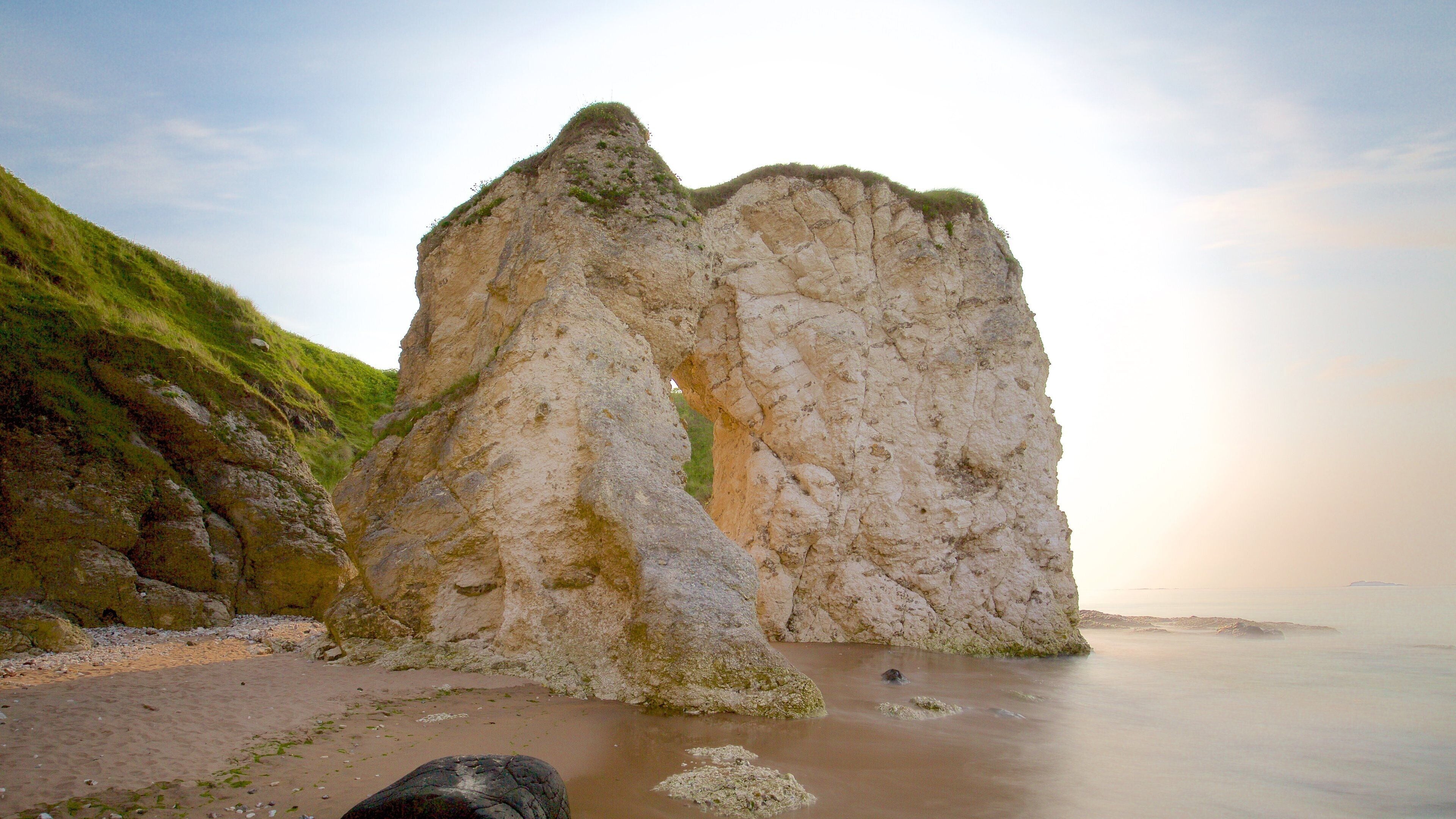 A large rock formation with a narrow passageway, surrounded by grassy hills and a calm sea.