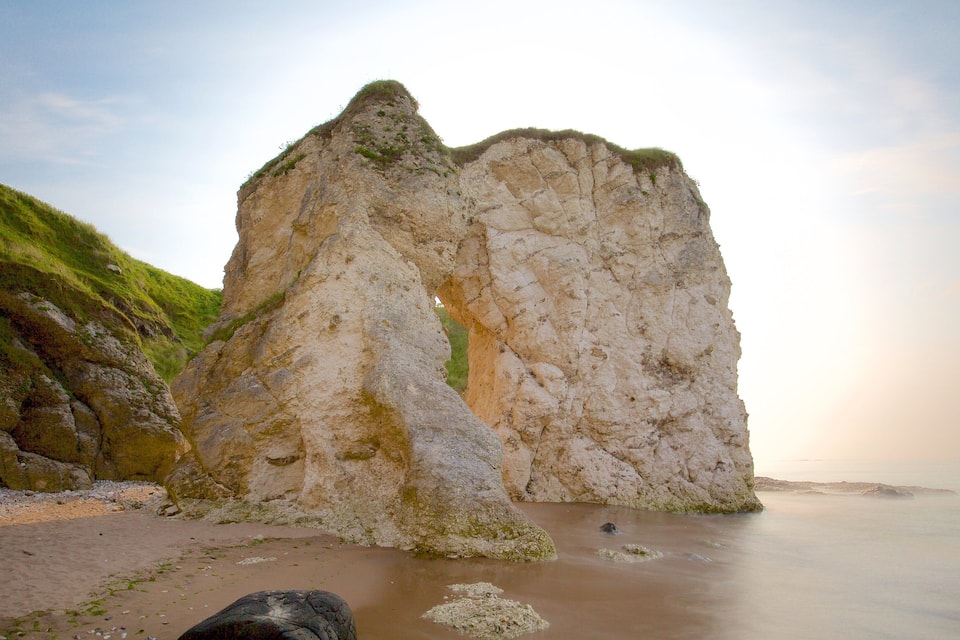 A large rock formation with a narrow passageway, surrounded by grassy hills and a calm sea.