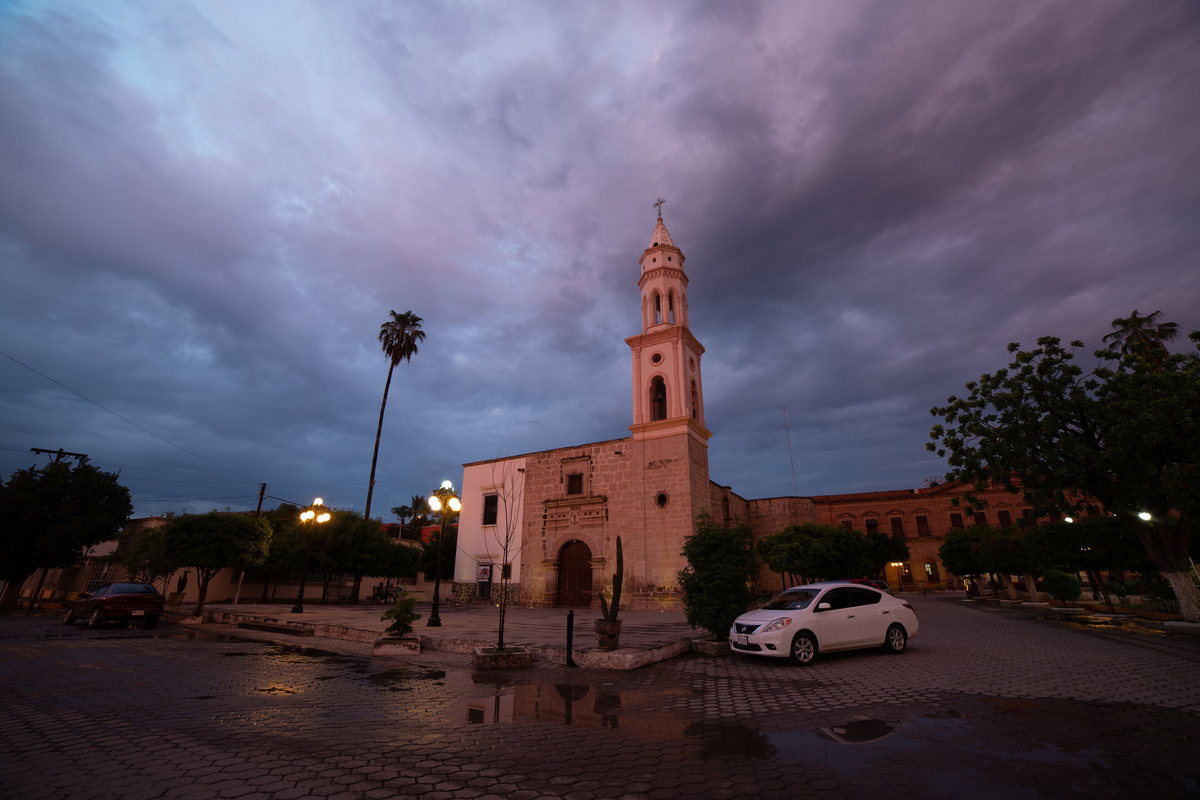 A historic church with a bell tower, surrounded by palm trees and a cobblestone street.