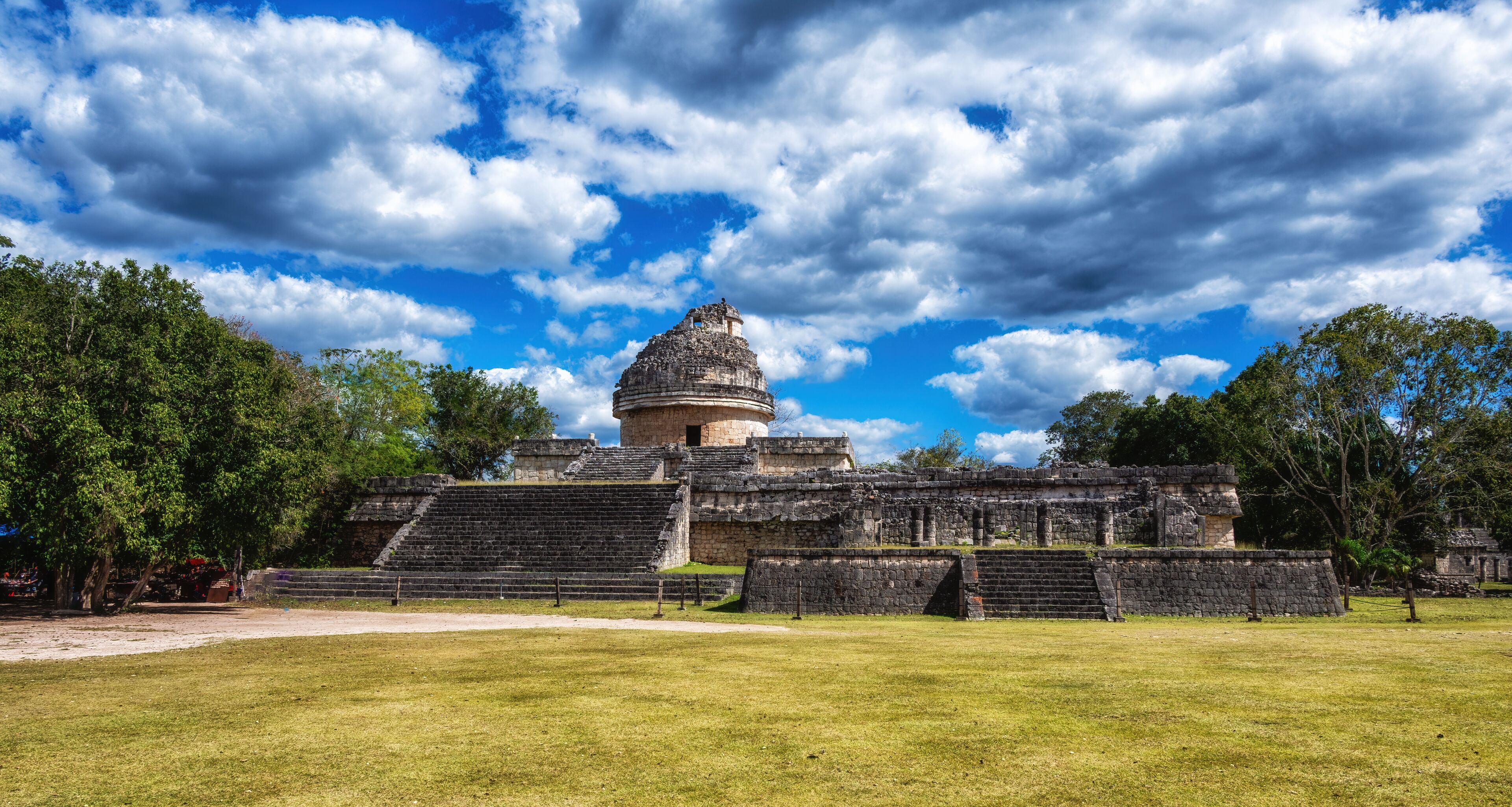 A historical stone structure with a large dome and steps, surrounded by a grassy field and trees under a partly cloudy sky.