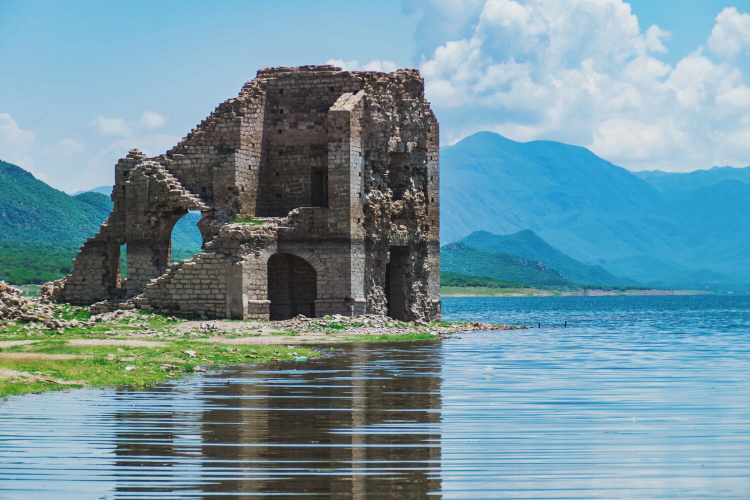 A ruined stone structure by a lake with mountains in the background.