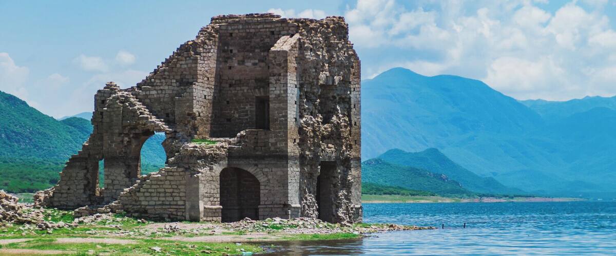 A ruined stone structure by a lake with mountains in the background.