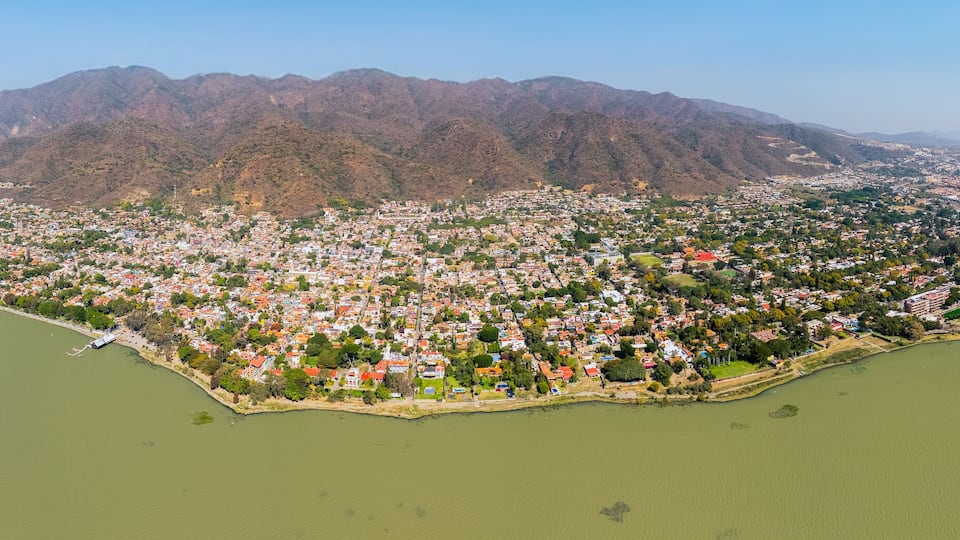 An aerial view of a coastal city with a dense urban area, a large body of water, and a mountain range in the background.