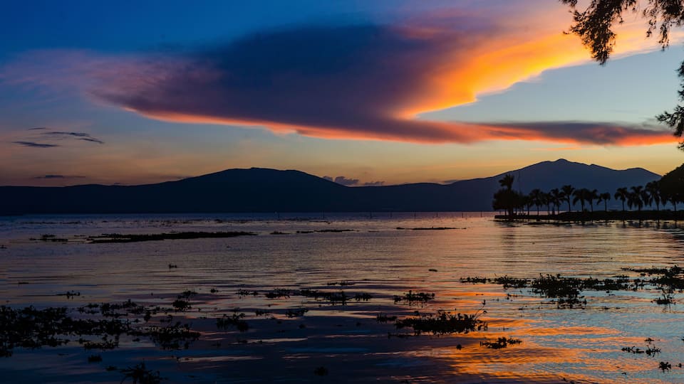 A sunset over a calm lake with silhouettes of mountains and palm trees.