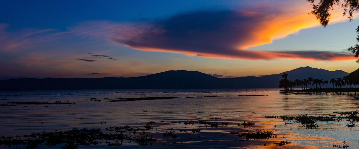 A sunset over a calm lake with silhouettes of mountains and palm trees.