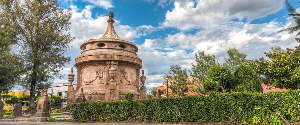 A historic stone monument with intricate carvings, surrounded by a well-maintained hedge and trees.