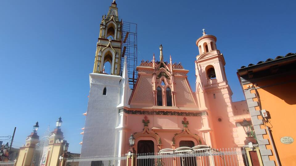 A pink and white church with a bell tower and a smaller tower, surrounded by a white fence.