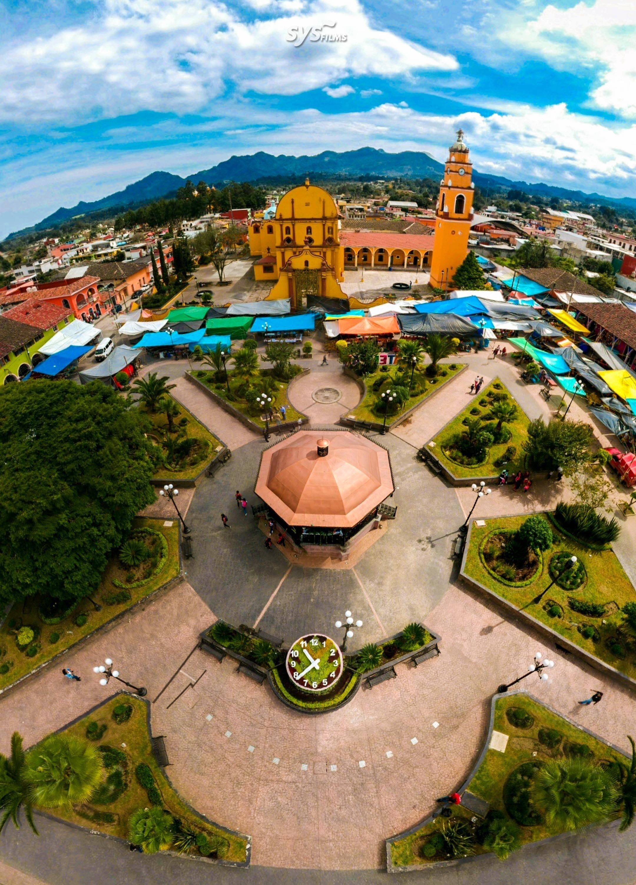 A town square with a central clock, surrounded by colorful market stalls and a prominent yellow church with a bell tower.