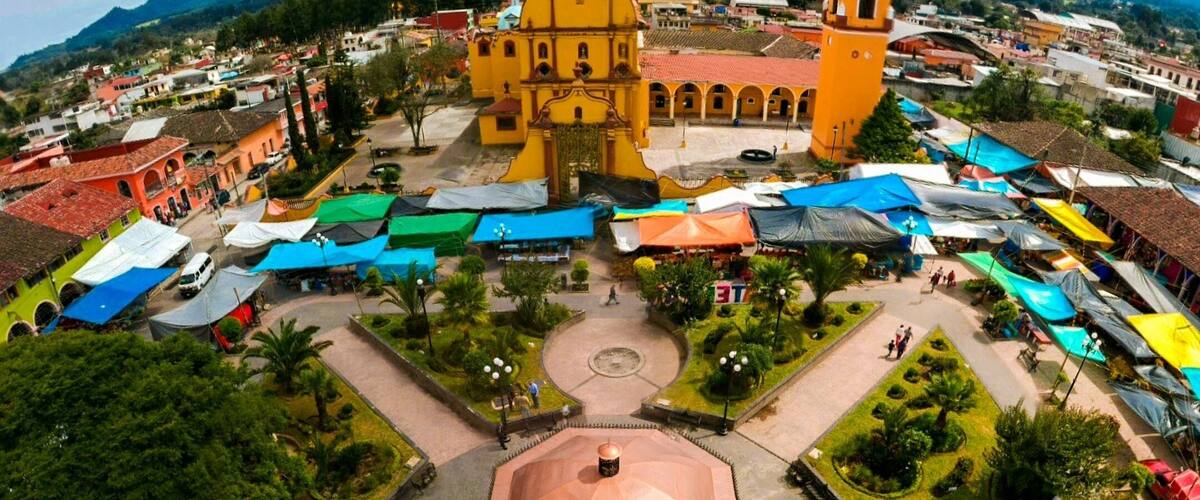 A town square with a central clock, surrounded by colorful market stalls and a prominent yellow church with a bell tower.