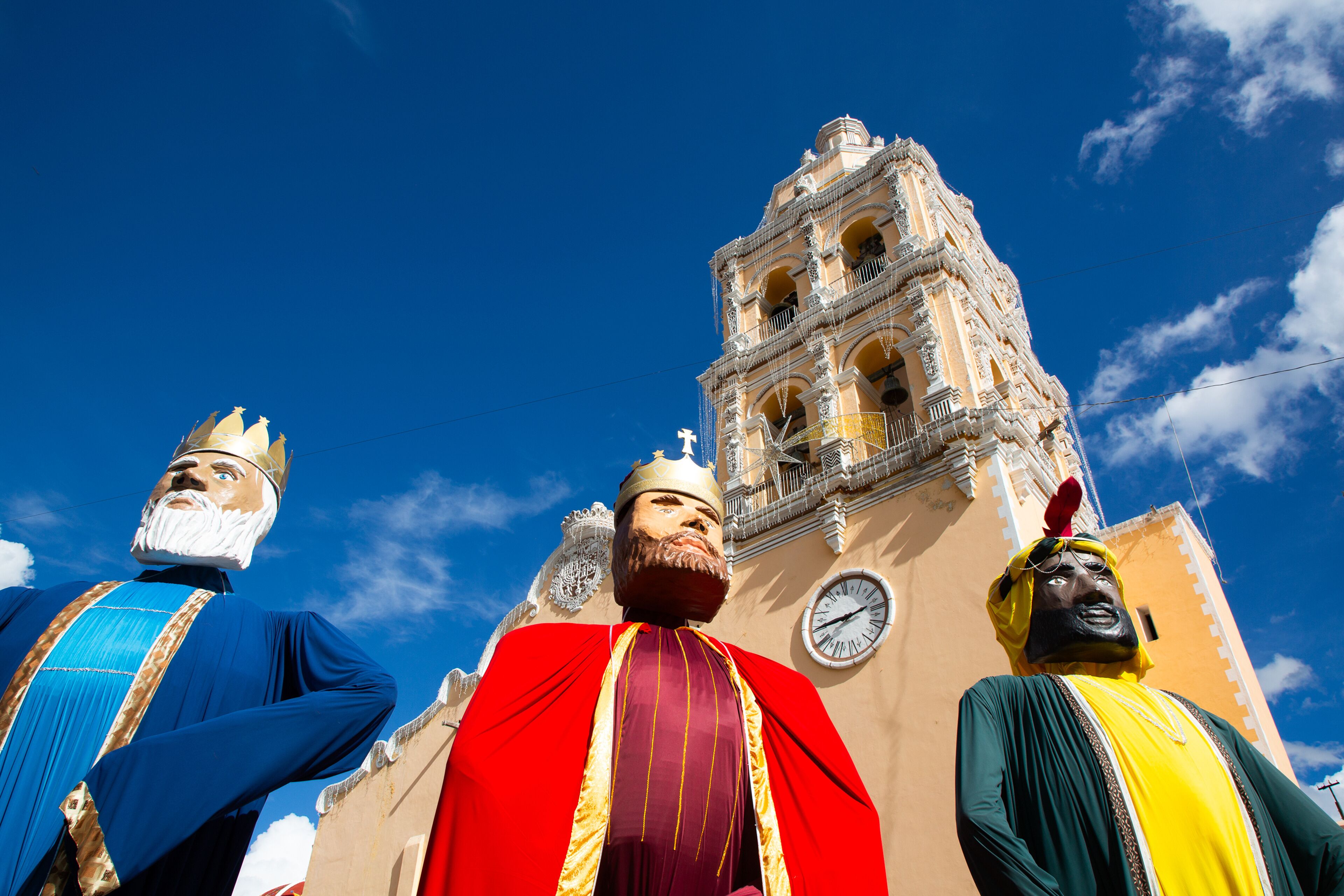 A large statue of a figure in a blue robe with a crown, a figure in a red robe with a crown, and a figure in a green robe with a crown, standing in front of a church with a clock tower.