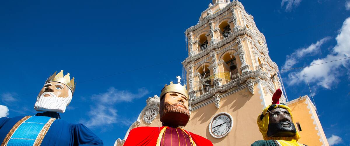 A large statue of a figure in a blue robe with a crown, a figure in a red robe with a crown, and a figure in a green robe with a crown, standing in front of a church with a clock tower.