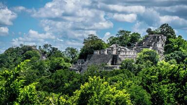 A Mayan pyramid surrounded by dense green foliage under a partly cloudy sky.