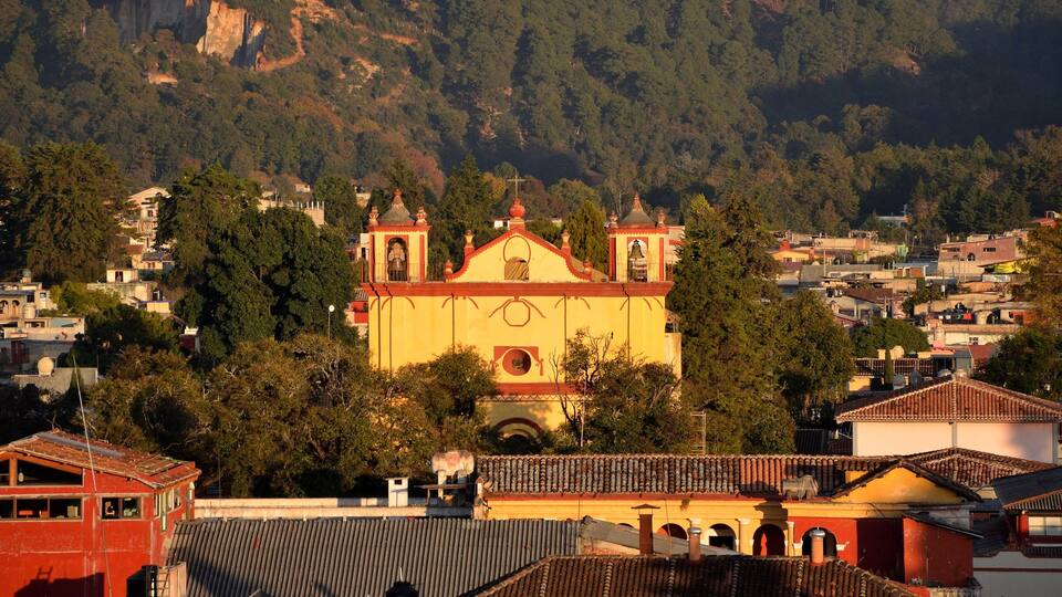A church with a bell tower surrounded by residential buildings and dense forested hills.