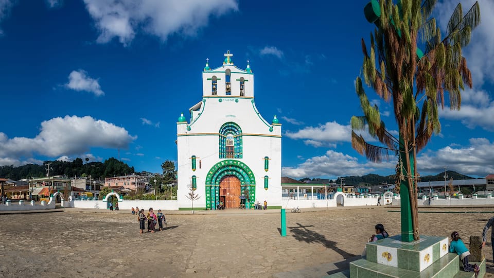 A white church with green accents and a wooden door, surrounded by a plaza with people and a palm tree.