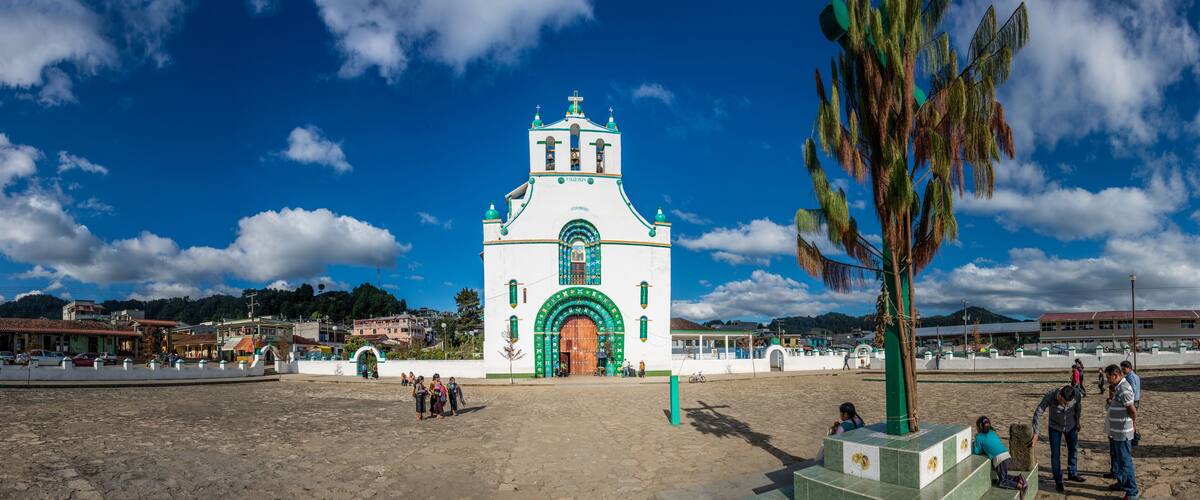 A white church with green accents and a wooden door, surrounded by a plaza with people and a palm tree.