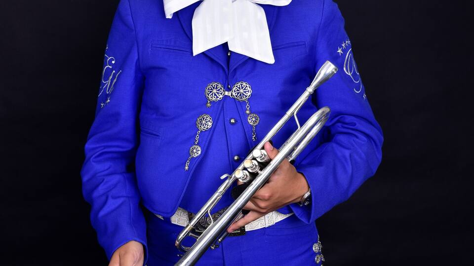 A person in a blue suit with a large sombrero, holding a silver trumpet.