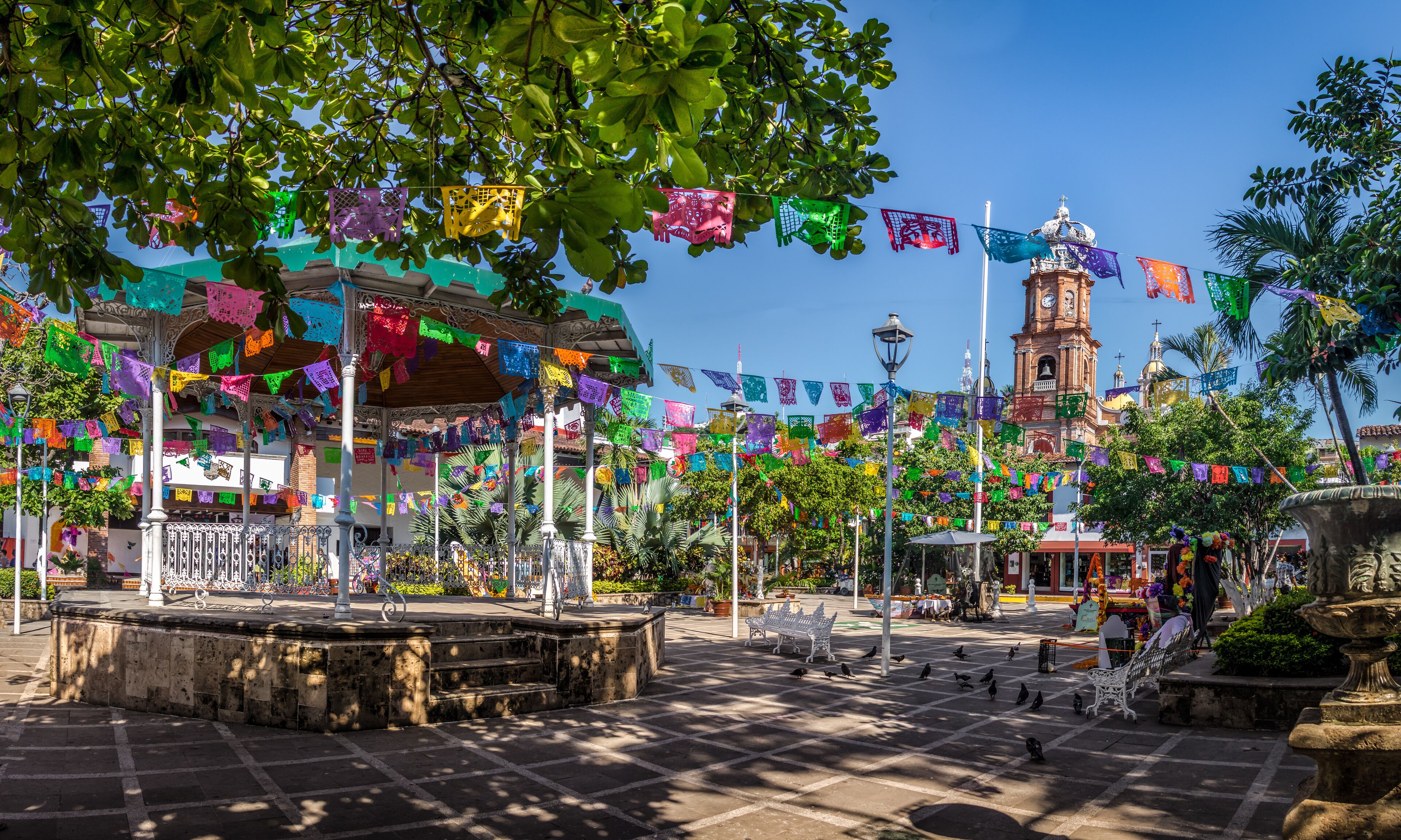 A plaza with colorful papel picado banners, a historic clock tower, and a fountain.
