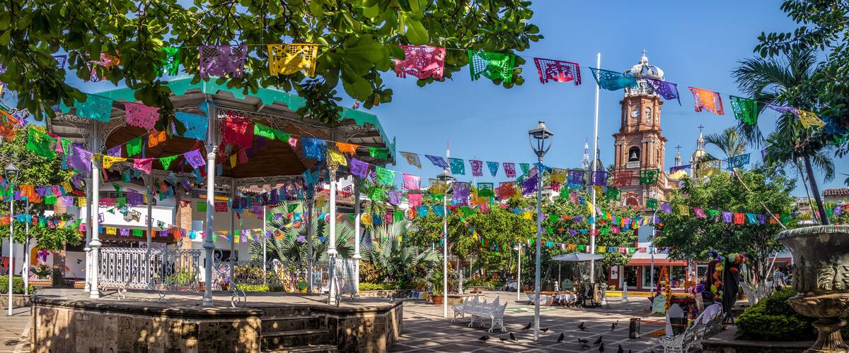 A plaza with colorful papel picado banners, a historic clock tower, and a fountain.