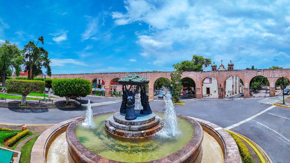 A round fountain with statues, a brick bridge, and a clear blue sky.