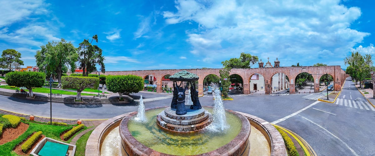 A round fountain with statues, a brick bridge, and a clear blue sky.