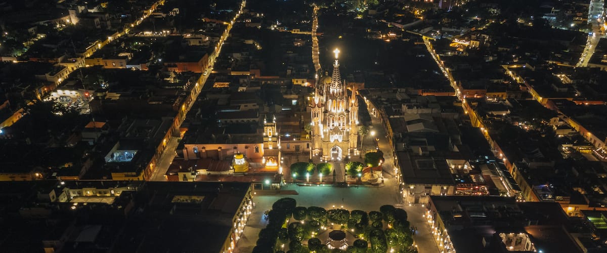 An aerial view of a city at night with illuminated streets, a prominent cathedral, and a well-lit square with trees.