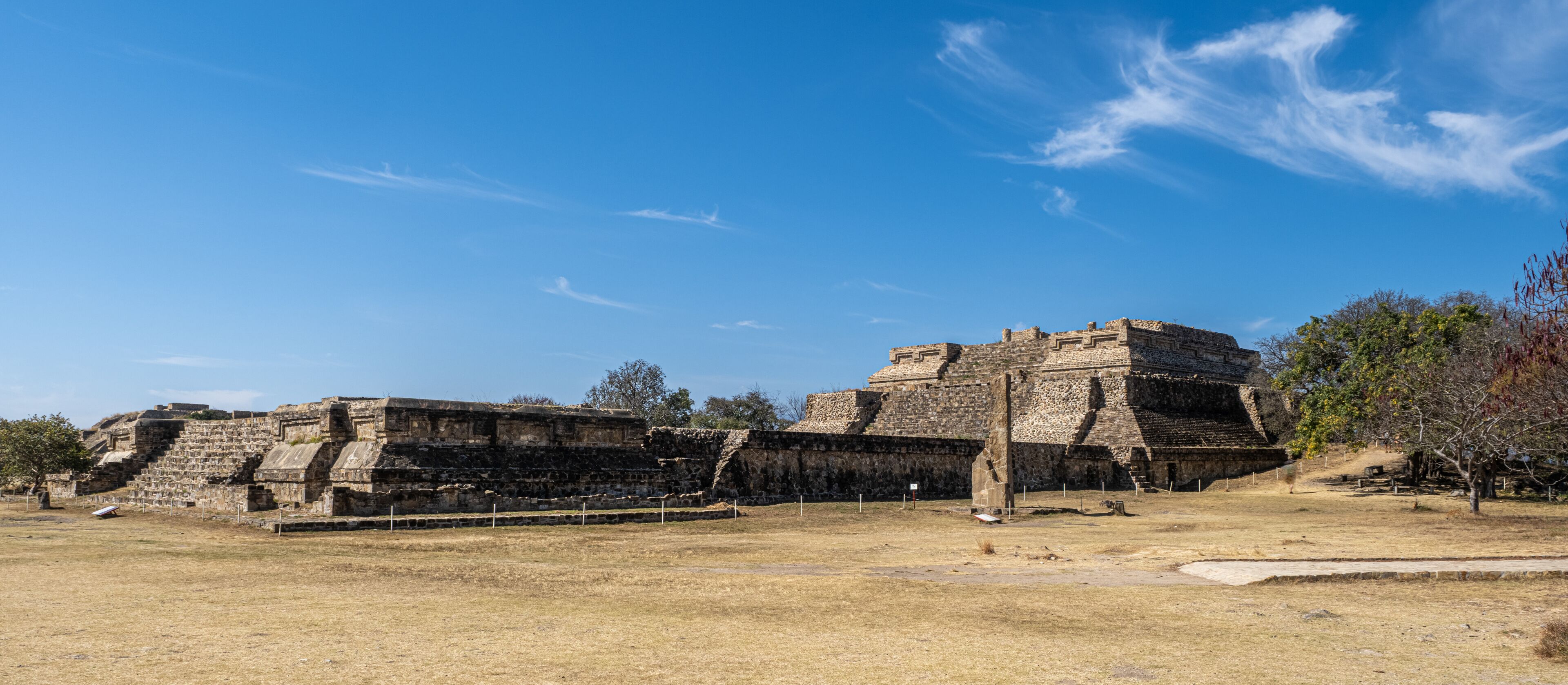 An ancient stone pyramid structure with a flat top and a stepped base, surrounded by dry grassland and trees.