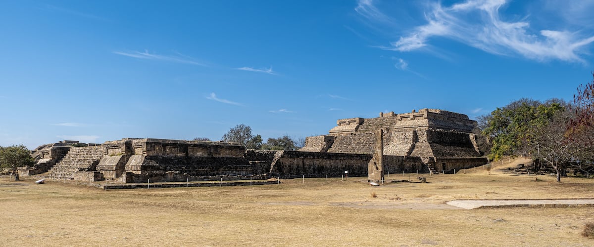 An ancient stone pyramid structure with a flat top and a stepped base, surrounded by dry grassland and trees.