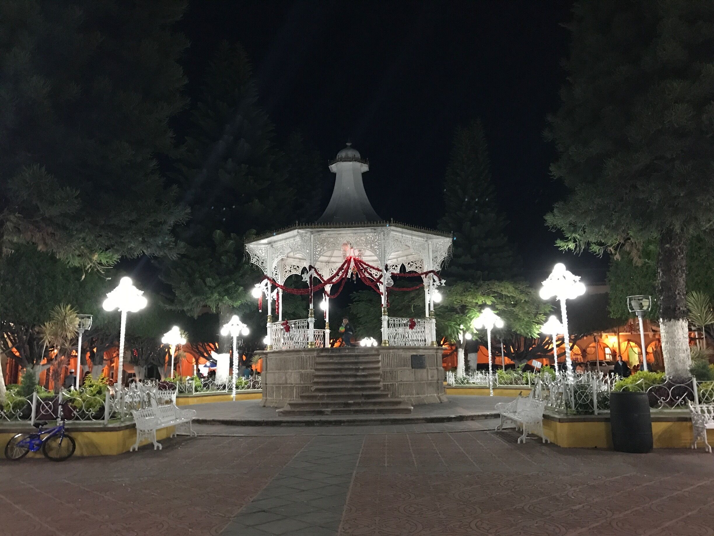 A gazebo with ornate railings and a dome roof, surrounded by trees and illuminated by street lamps.