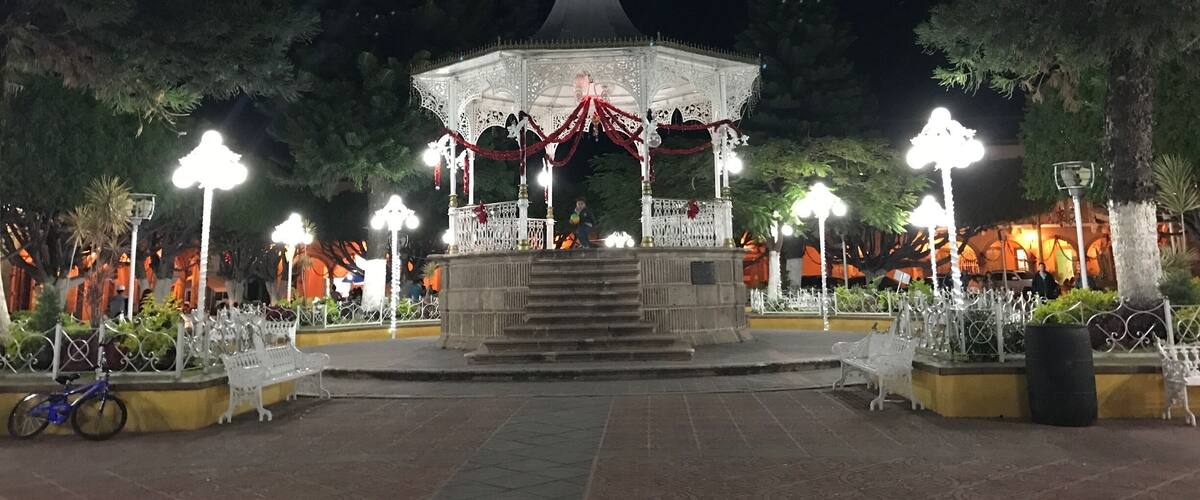A gazebo with ornate railings and a dome roof, surrounded by trees and illuminated by street lamps.