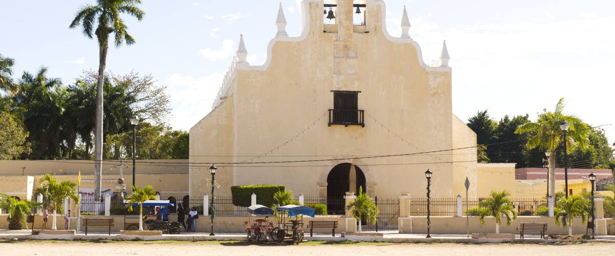A colonial-style building with a bell tower, palm trees, and a paved plaza.