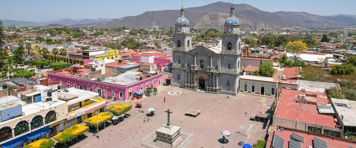 A plaza with a church, colorful buildings, and mountains in the background.