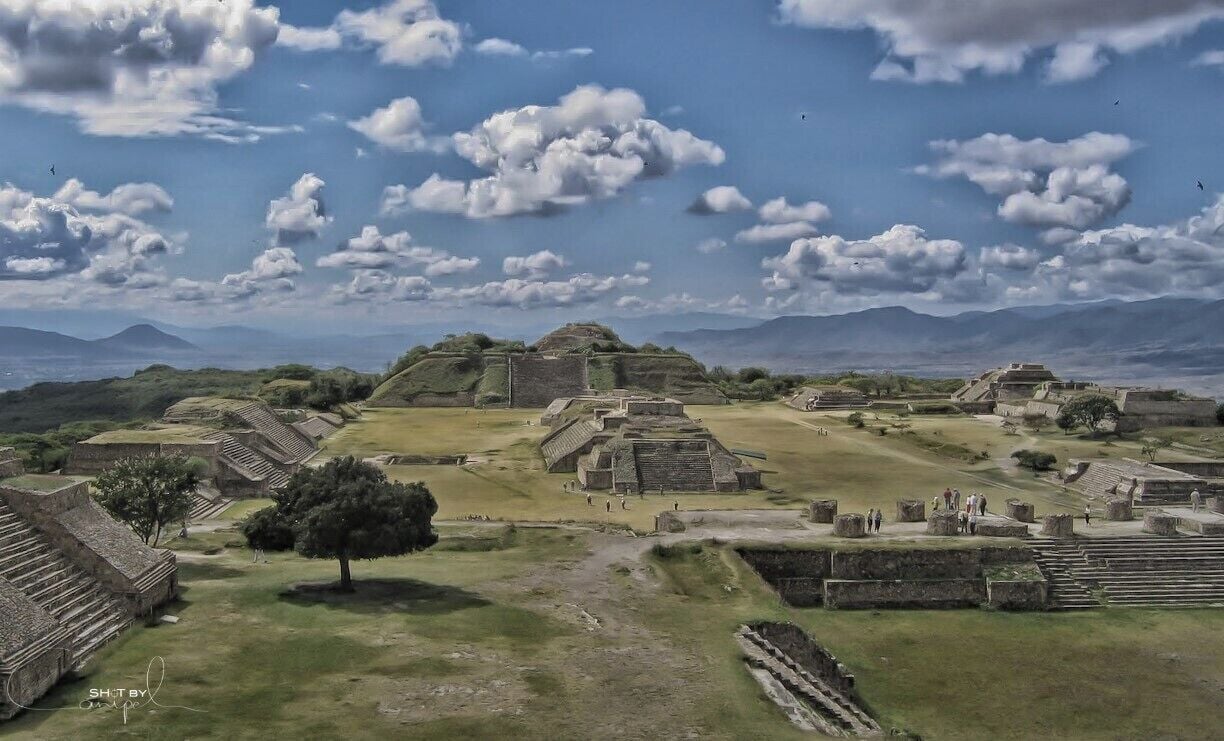 Ancient ruins with stepped pyramids and a large central structure, surrounded by open fields and distant mountains.