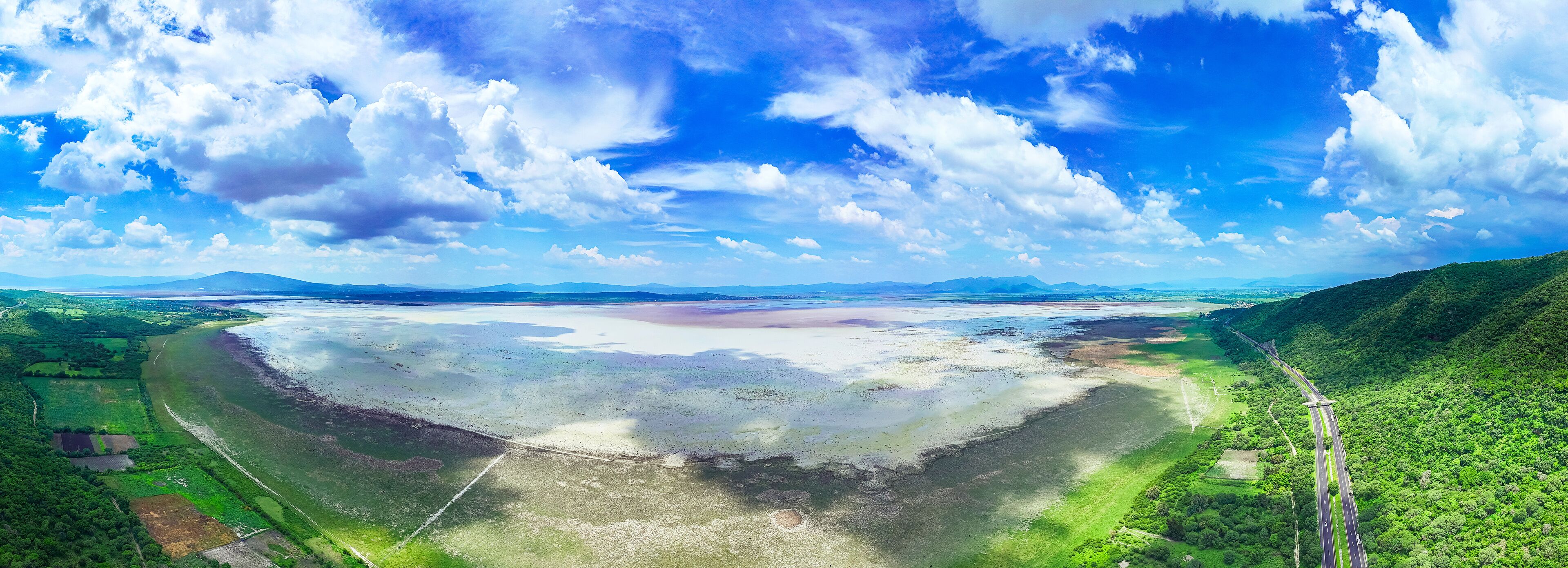 A coastal landscape with a highway curving through green hills, a large body of water, and distant mountains under a partly cloudy sky.