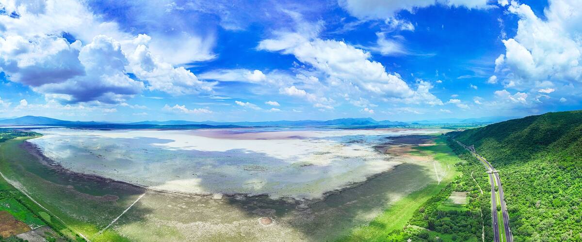 A coastal landscape with a highway curving through green hills, a large body of water, and distant mountains under a partly cloudy sky.